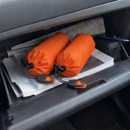 PACK 2: dos sacos vivac térmicos guardados en sus fundas naranjas, cada uno con su silbato de rescate, dentro de la guantera de un coche