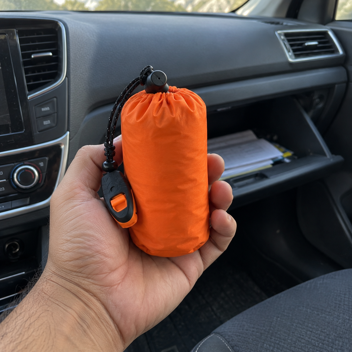Saco vivac térmico guardado en su funda, sostenido en la palma de la mano dentro de un coche